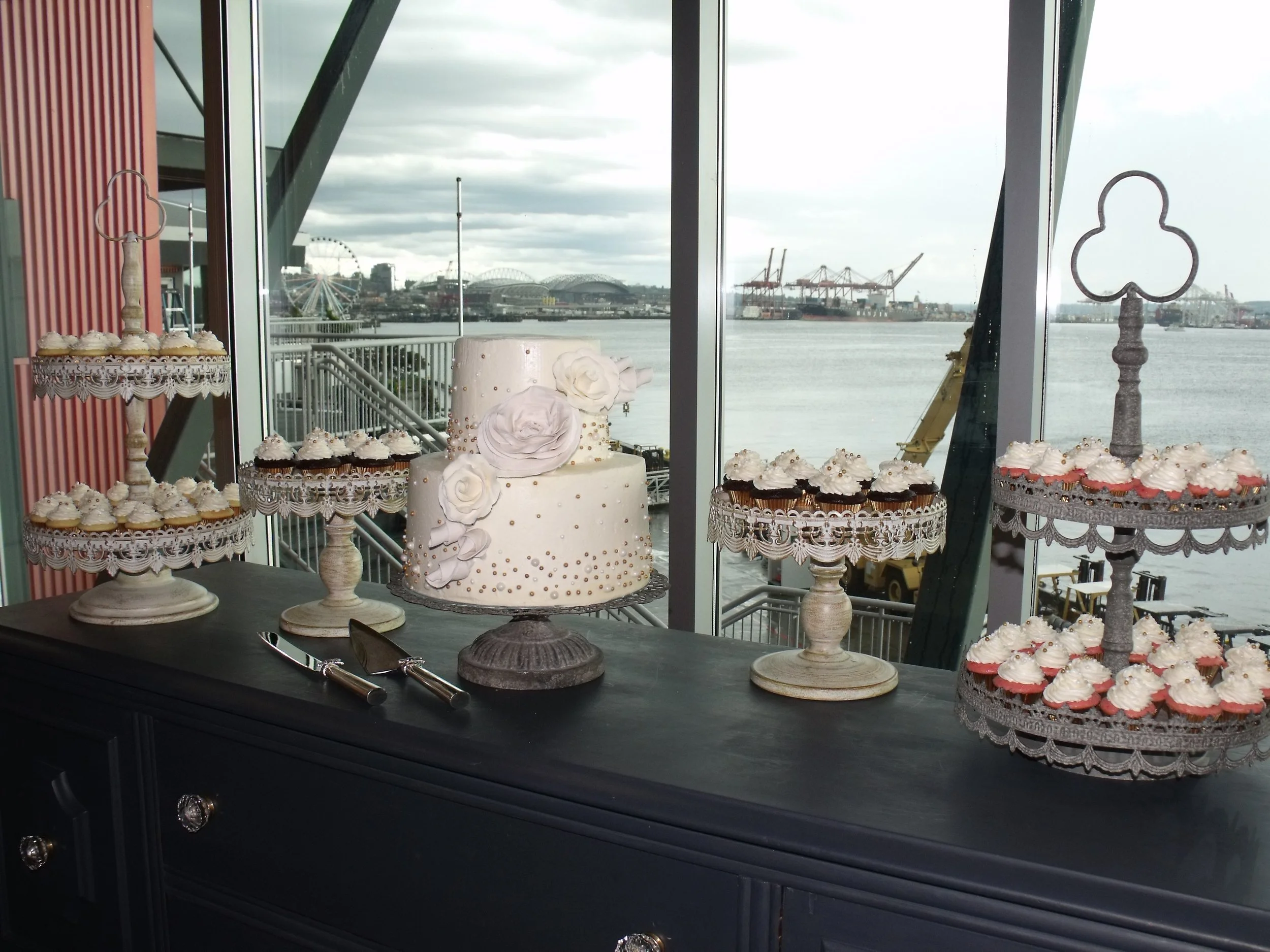 Cake and Cupcake Wedding Two Tier Stacked Wedding Cake on Uplifted Gray Cake Plate White and Gold Sugar Pearl design Large Floribunda White Sugar Roses and Buds Four other Cake Plates  with Mini Cupcakes 