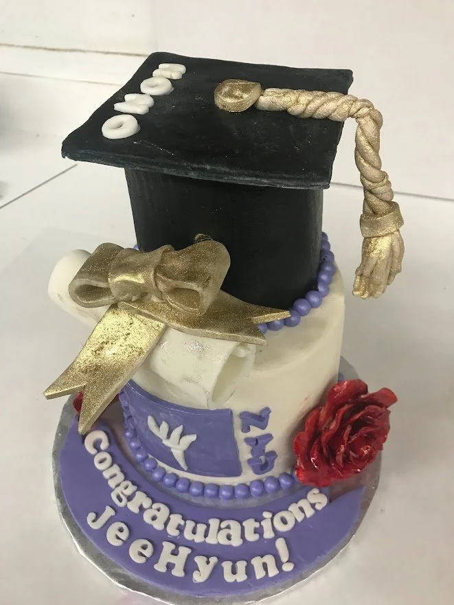 Small Two Tier Round Buttercream Frosted Graduation Cake Top Cake Designed as Traditional Graduation Cap with Fondant Mortar Board Tassel Year White Fondant Scroll and Ribbon Sugar Metallic Over Brushing of Ribbon and Tassel Large Red Floribunda Rose
