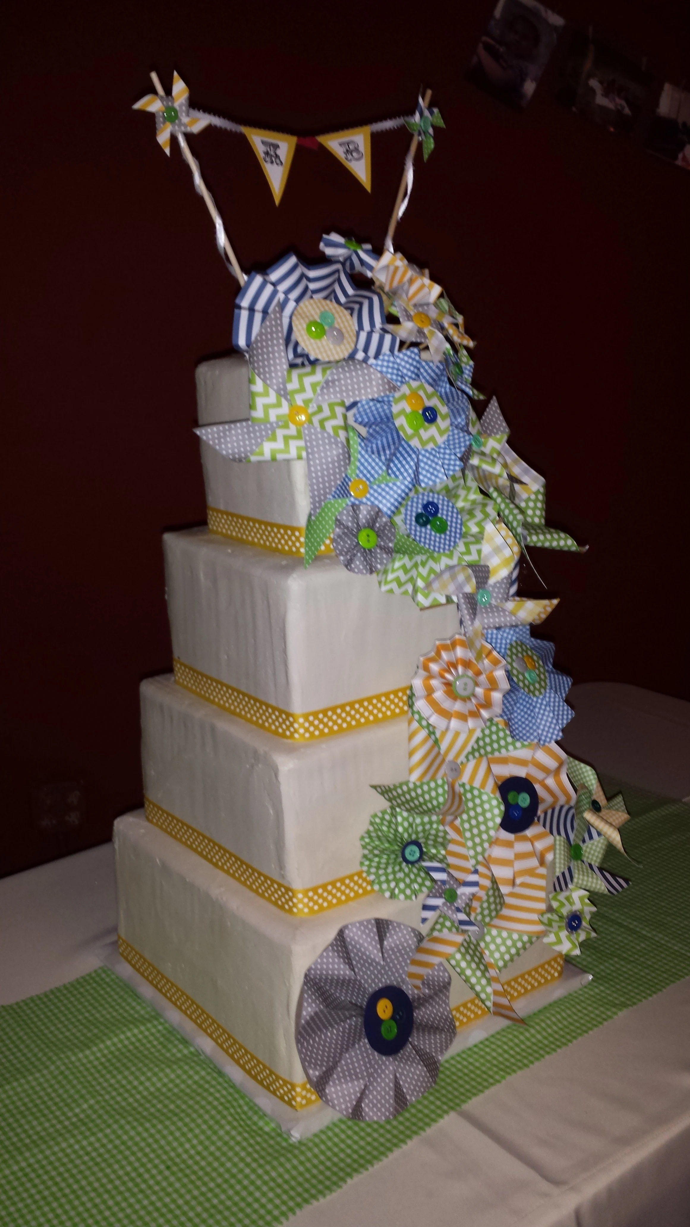 A four-tiered white cake decorated with yellow and white polka dot ribbon, topped with a cascade of colorful paper pinwheels and paper flowers in shades of yellow, blue, gray, and green with button centers, and a small banner with flags on top.