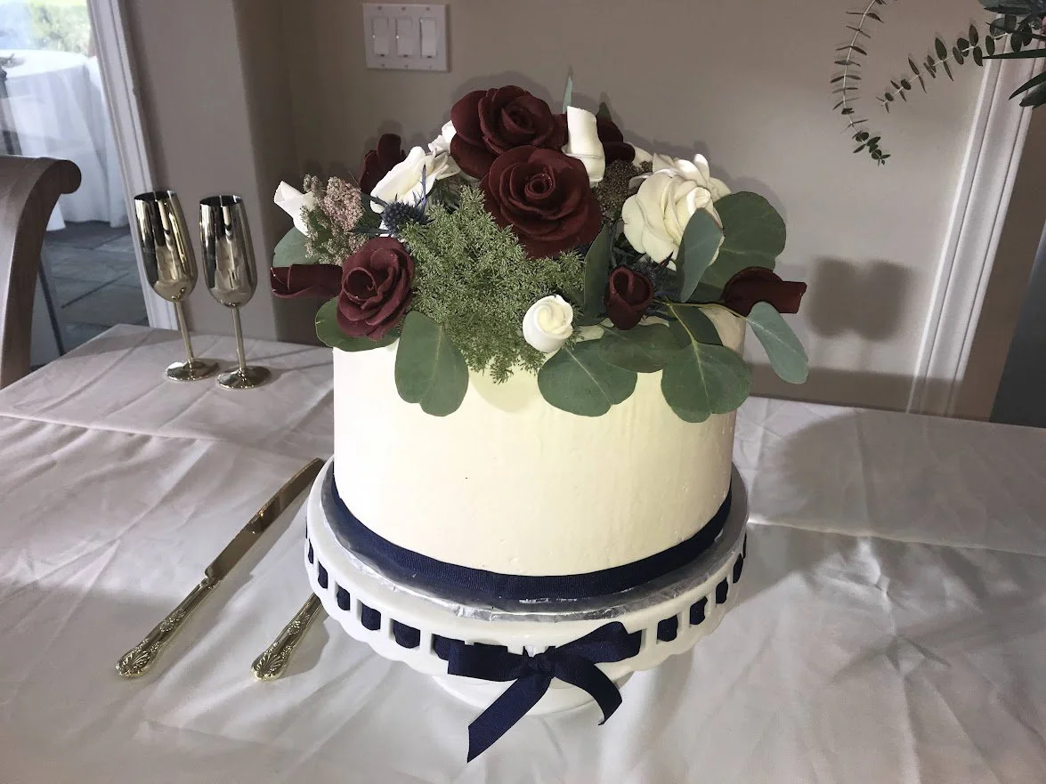 Small Round Wedding Celebration Cake on Whiter Pillar Plate Navy Blue Cross grain Ribbon Buttercream Frosted Fresh Greens Sugar Porcelain Burgundy Red and White Roses Navy Fondant Bottom Border