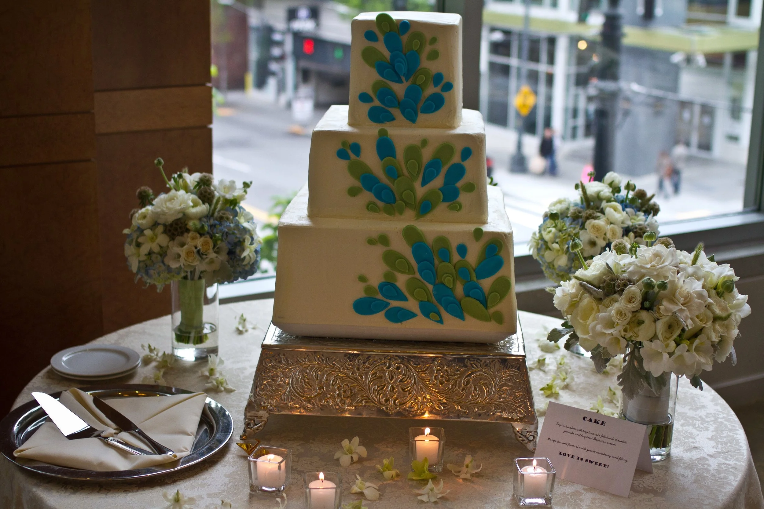 A three-tier wedding cake decorated with blue and green fondant patterns, displayed on a table with floral arrangements and candles, near a window with an urban street view in the background.