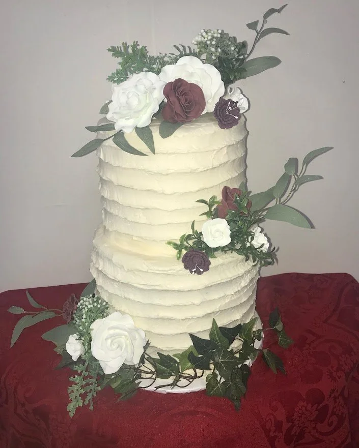Romantic Old Fashioned  Buttercream Troweled  Two Tier Stacked Wedding Cake Sugar Porcelain White Gardenias and Burgundy Full Open Roses Set Over Artificial and Fresh Greens Deep Red Table Cloth