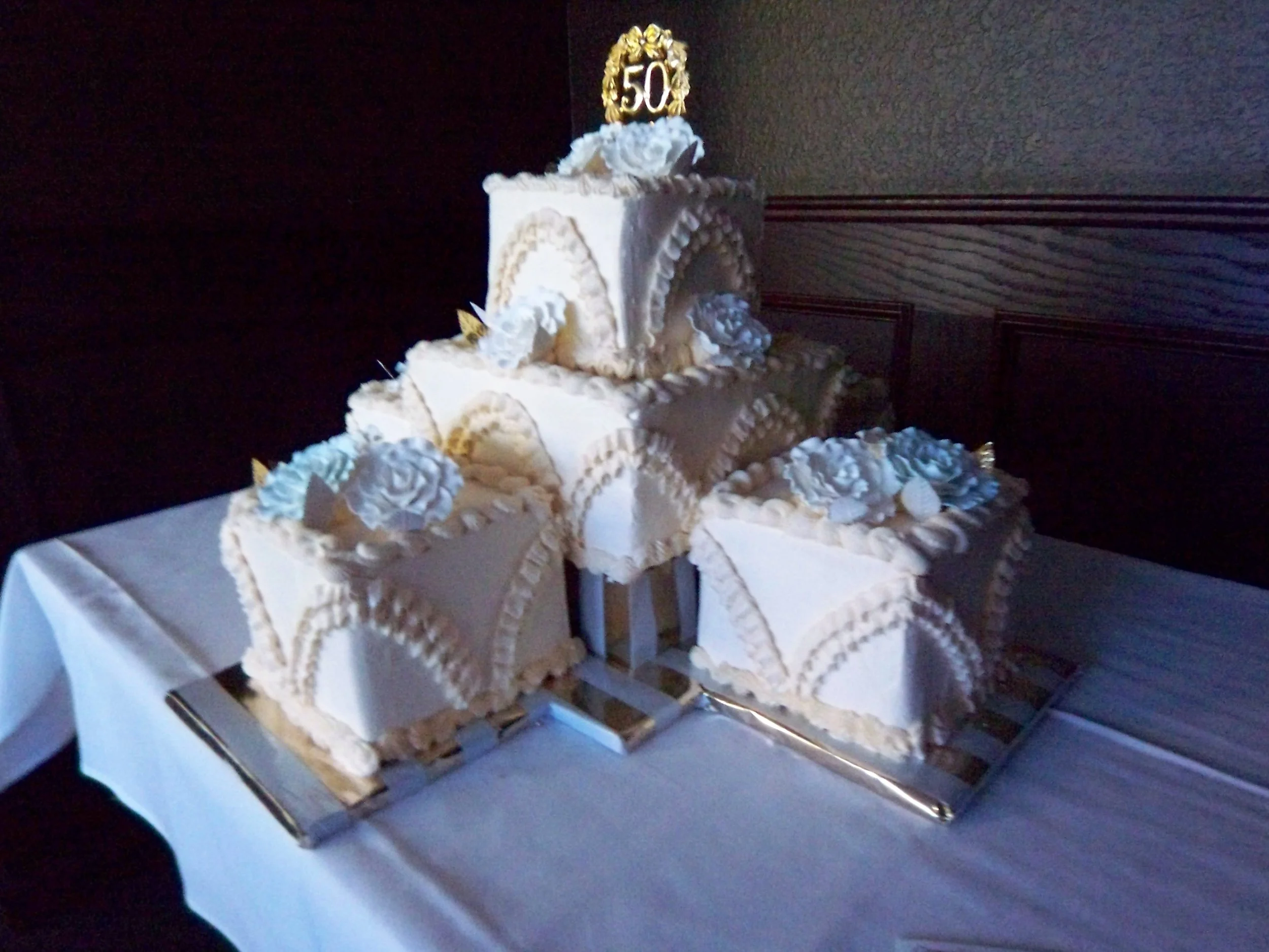 50th wedding anniversary cake four square buttercream frosted square two tier stacked cake two side cakes dummy platform L shaped arrangement blue sugar dusted full open roses gold 50 topper