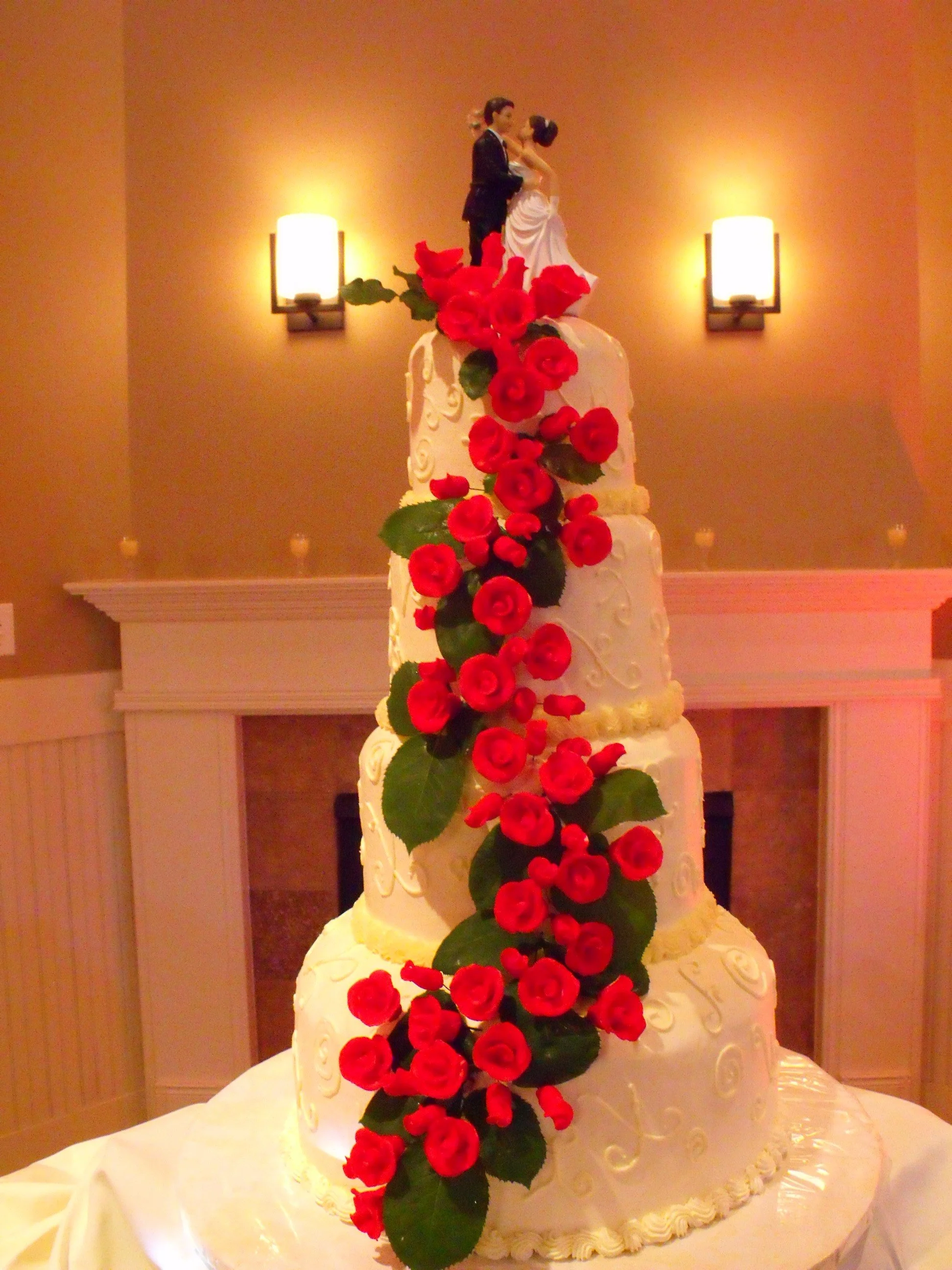 Four Tier buttercream frosted stacked rounds tower wedding cake piped scroll designs side of cakes flowing cascade red bud and open sugar porcelain roses over fresh greens backing bride and groom topper