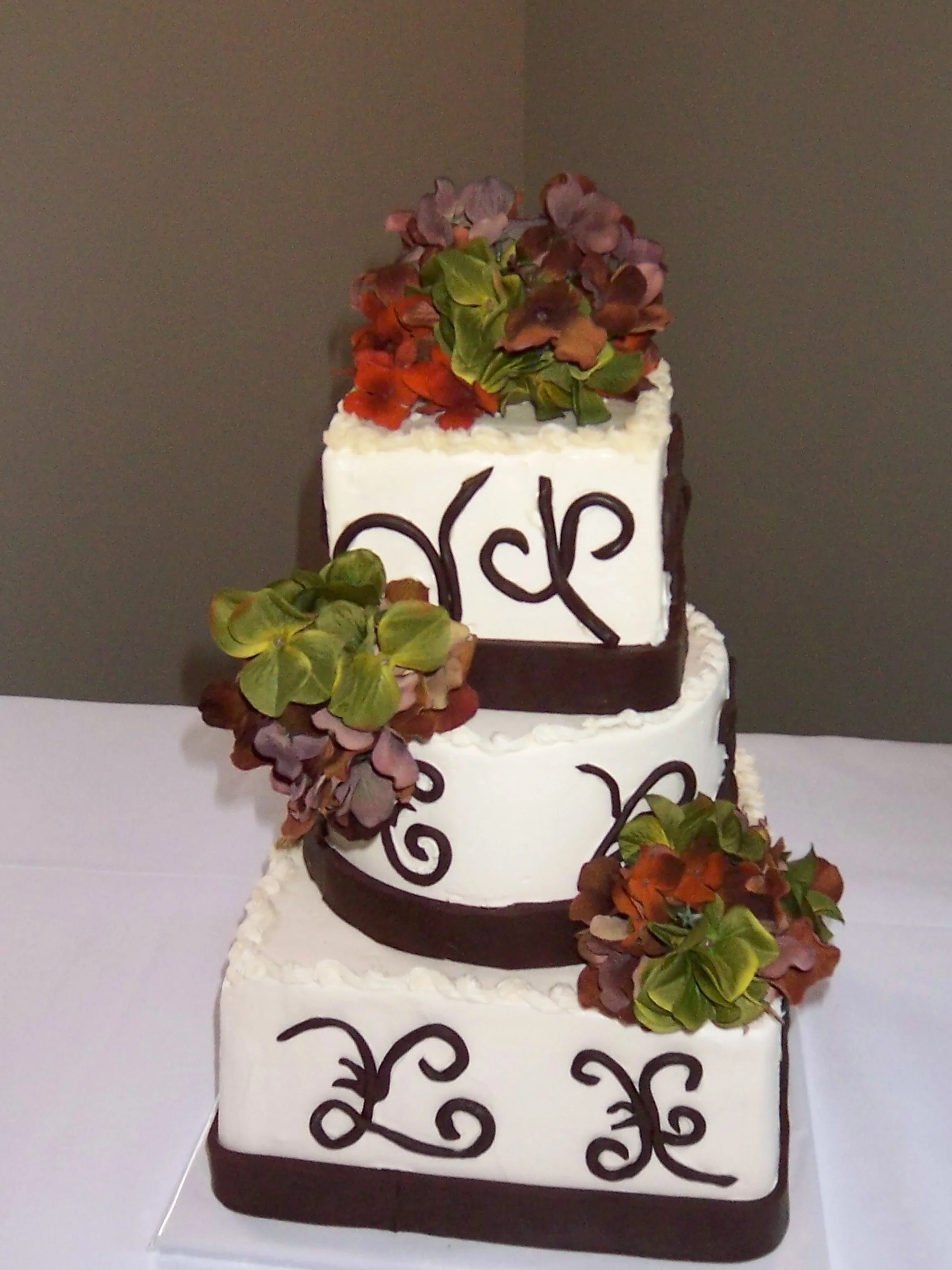 Wedding Cake with Square and Round Opposing Stacked Tiers Smooth Frosted Buttercream Tiers Banded with Chocolate Borders and Han Applied Chocolate Scroll Designs Three Autumn Silk Flower Poms