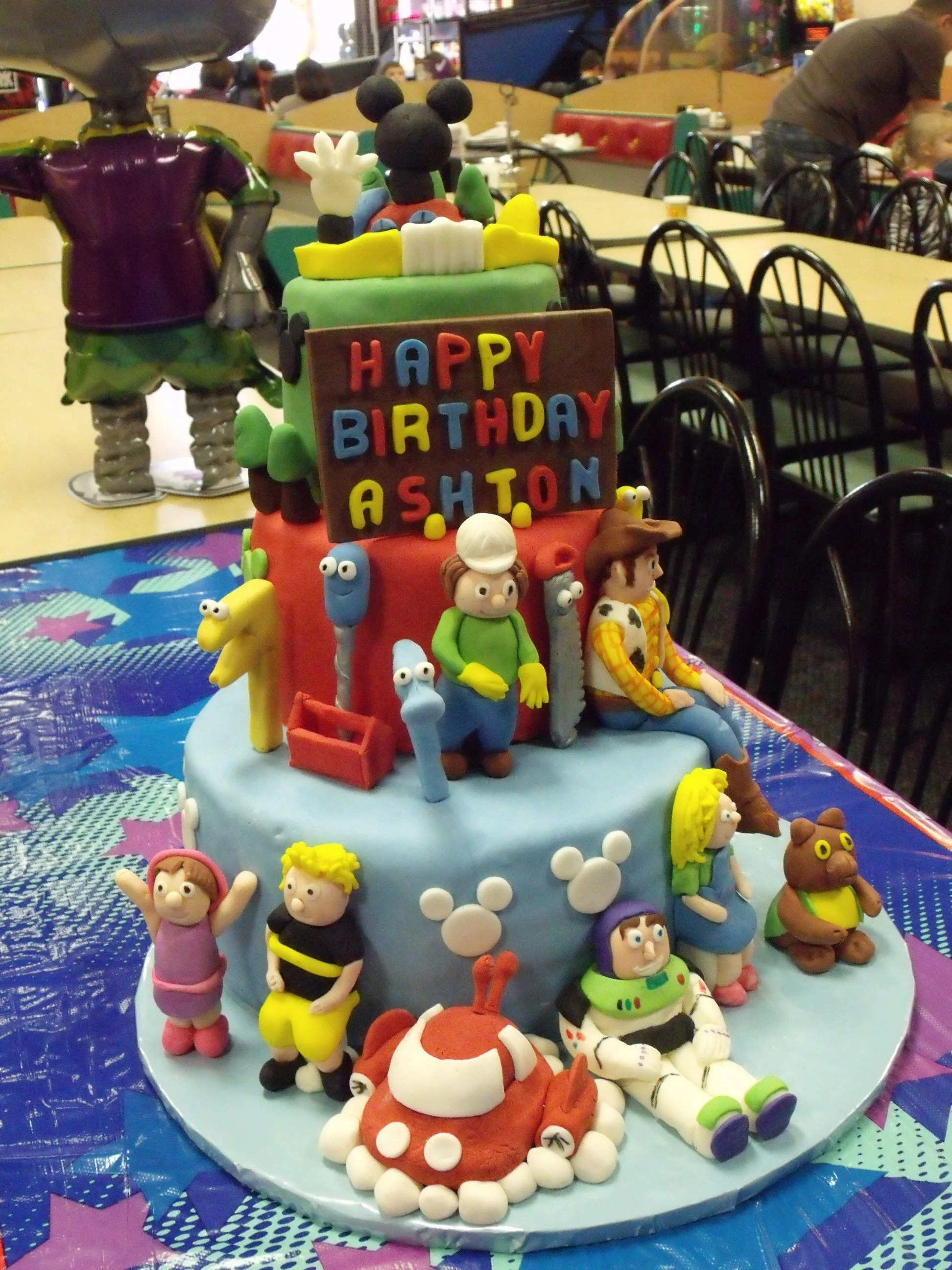 Birthday Cake Colorful Three Tier Fondant Covered Cake Fondant Creations of Mickeys Playhouse Buzz Lightyear and Space Ship and Woody Bob The Builder with  Friends and Salutation Billboard and Lettering Sky Blue Wrapped Board