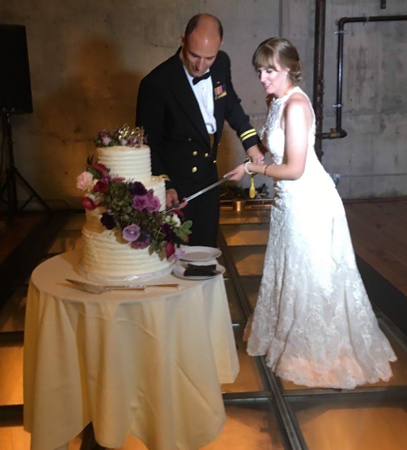 Large three tier stacked round Wedding Cake French buttercream frosted cake  sugar porcelain open pink purple lavender roses flowing design over fresh greens military sword cake cutting Bride and Groom cake table linen plates