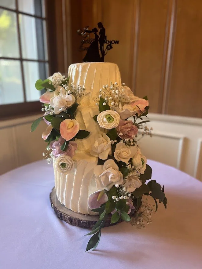 Three Tier Stacked Wedding Cake with Buttercream Diagonally Upswept Furls On Rustic Wood Round set with Sugar Porcelain Dusted White Peony's Gardenia's Rose's Pink throated Calla's on Live Greens and Baby's Breath Sprigs and Wrought Iron silhouette o