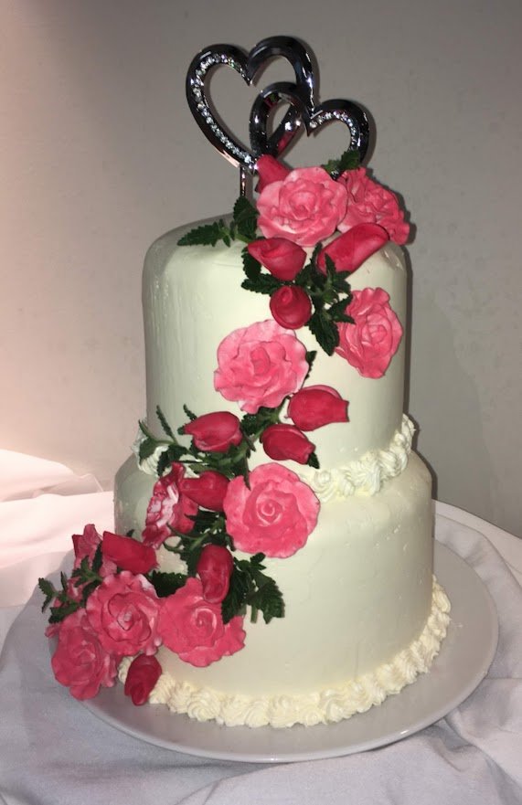 Romantic Two Stacked Tier Wedding Cake Frosted in Buttercream with Rounded Top Edge Fuchsia and Red Sugar Porcelain Roses over Fresh Greens with Silver Metallic Joined Hearts with Rhinestones Topper White Linen Table