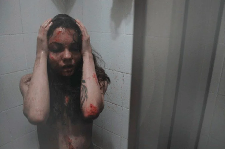 A woman with disheveled hair and makeup smeared on her face, appearing distressed, standing topless in a shower or tiled room with bloodstains on her skin and the wall.