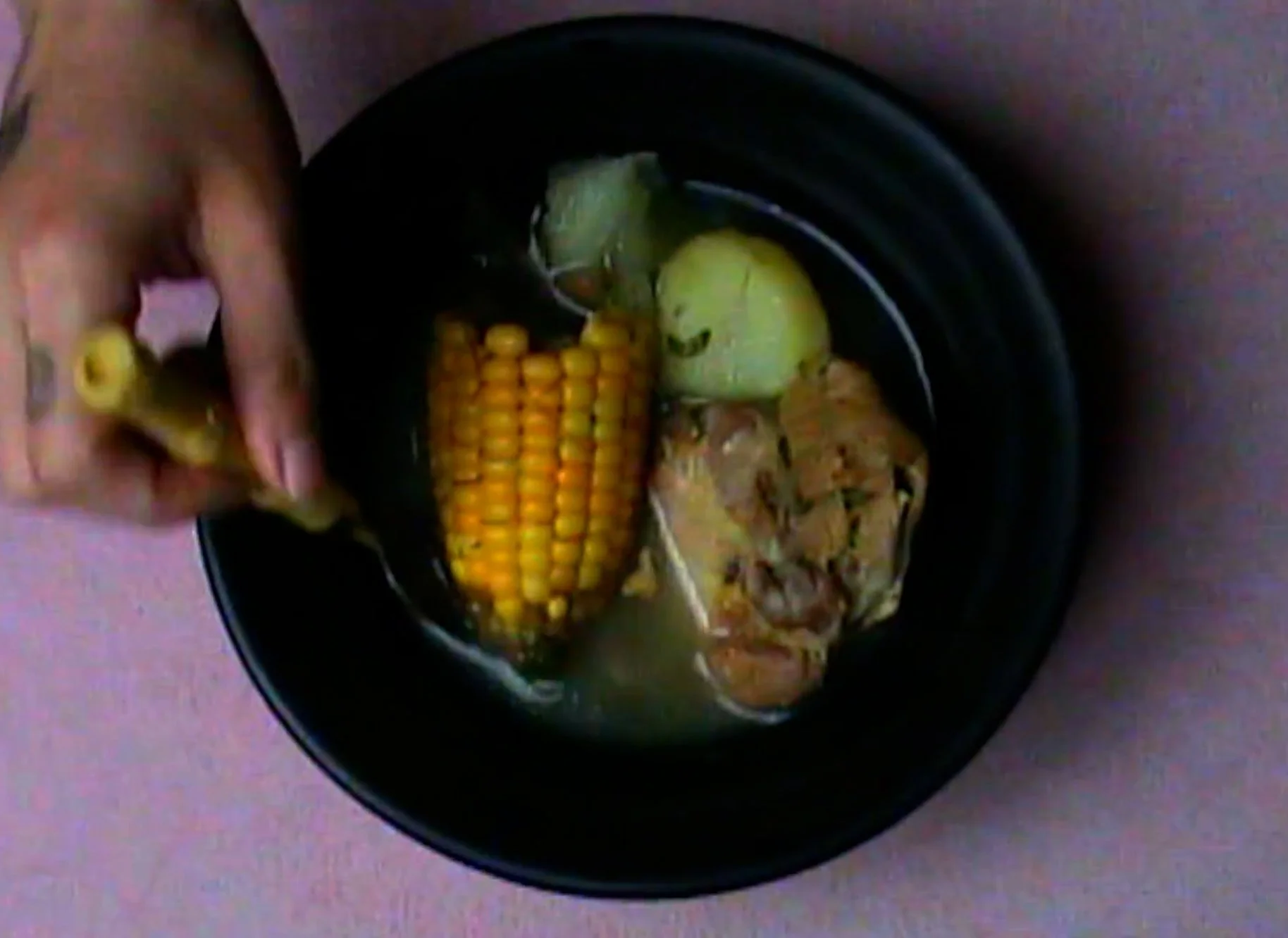 A black bowl with corn on the cob, pieces of cooked meat, and an apple in broth.