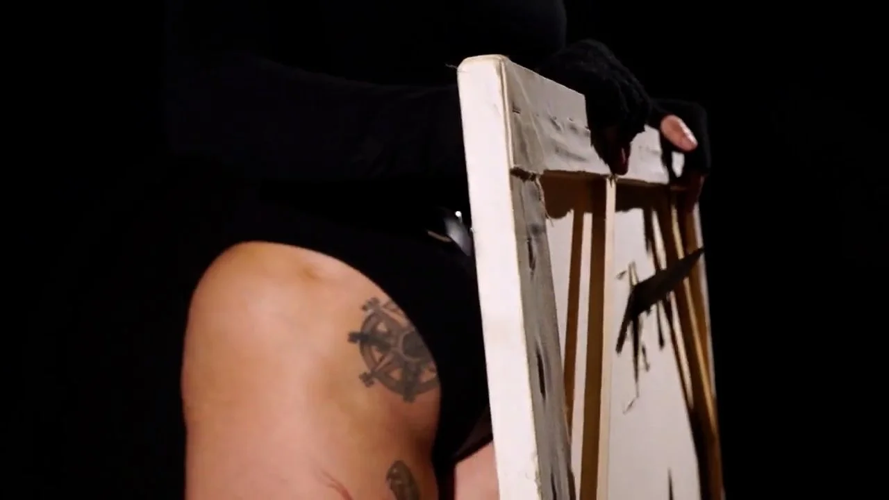 Person wearing black gloves holding a paintbrush, leaning over a wooden easel or palette on a black background, with visible tattoos on their arm.