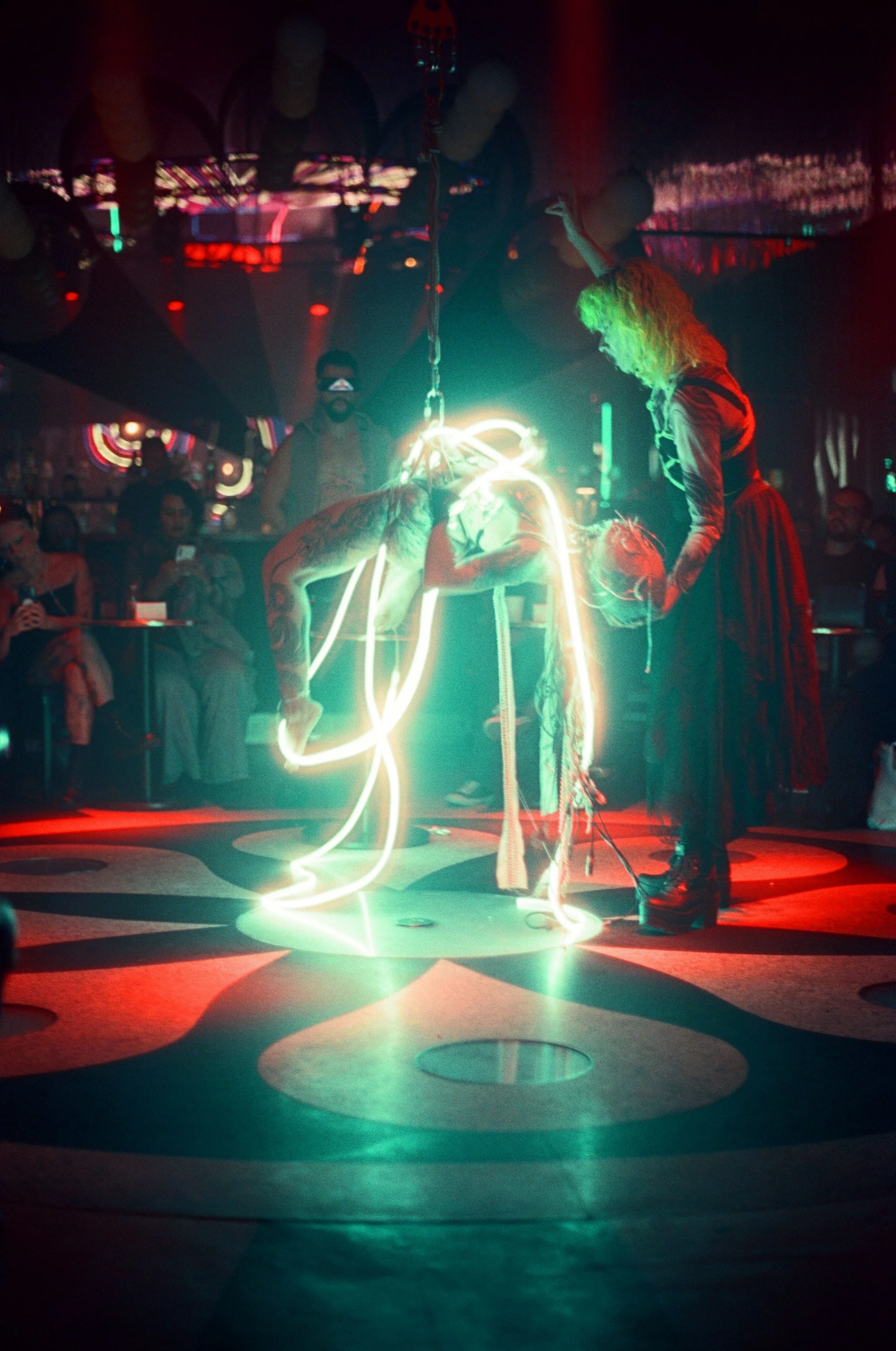 A performer doing an acrobatic pose illuminated by neon lights at a nightclub with audience watching.