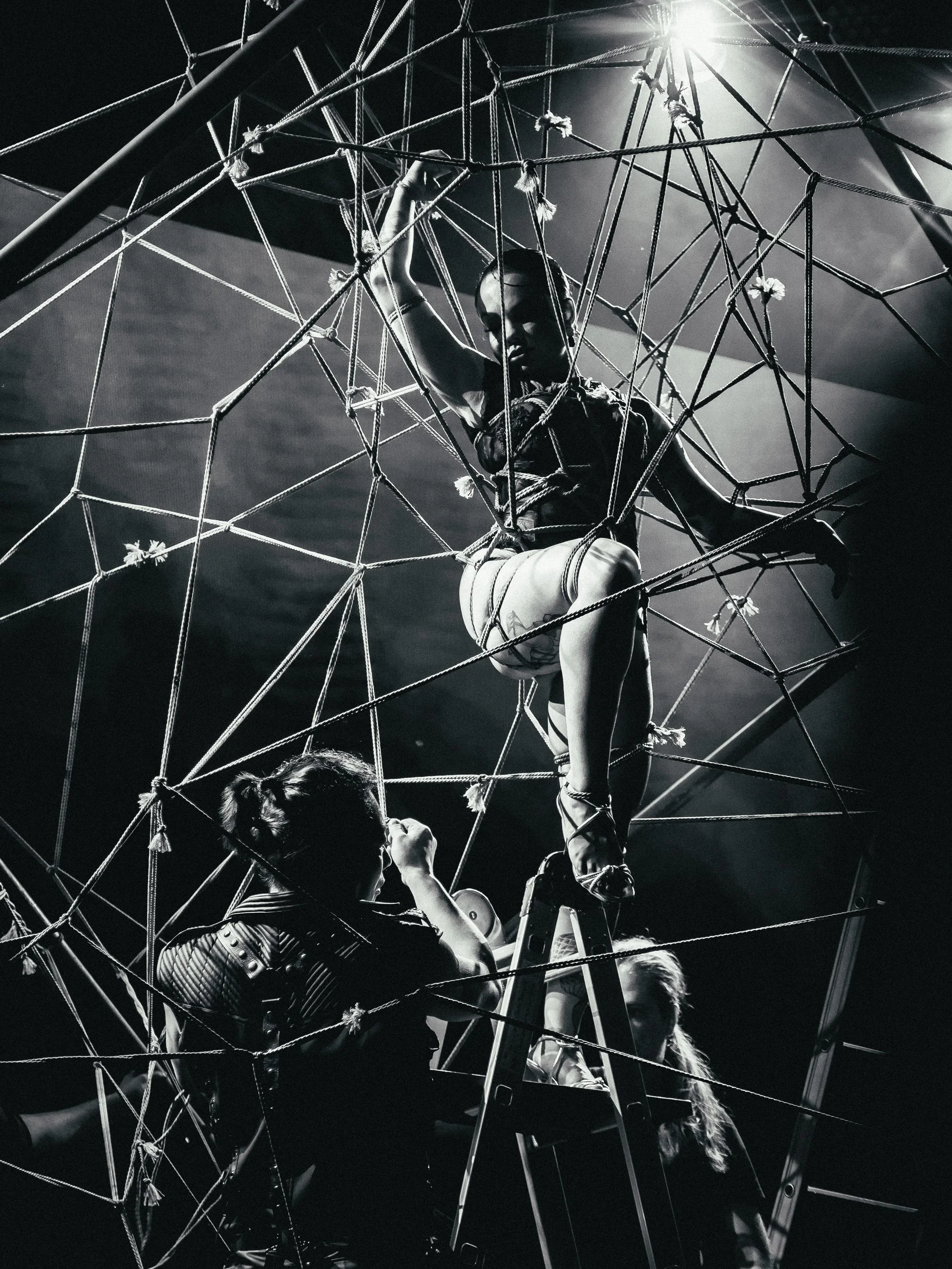 A woman climbing on a spherical structure made of ropes, with two other women on the ground, one on a ladder, as part of a performance or art piece in black and white.