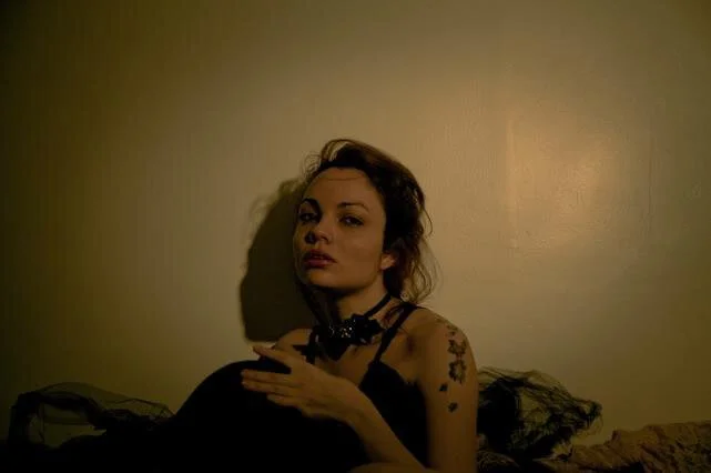 A woman with dark hair, wearing a black outfit and choker, sitting on a bed in a dimly lit room.