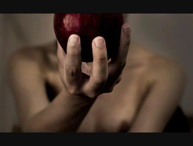 A person holding a red apple close to the camera, with their arm and shoulder visible in the background.