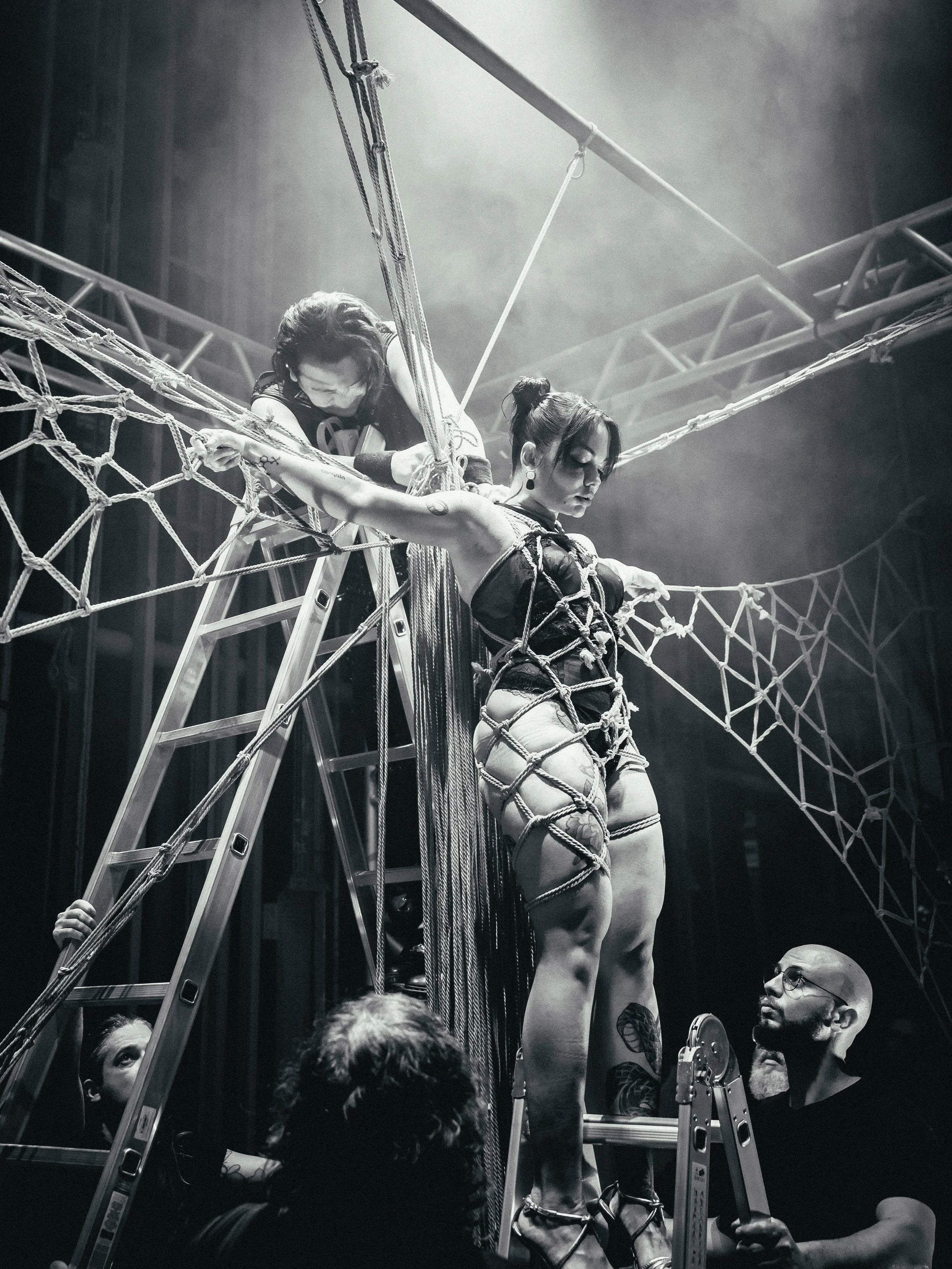 Two women on a stage with ropes and scaffolding, one standing and chained with ropes around her body, the other leaning over her, while a man holds a ladder and looks up at them, in a theatrical performance setting.