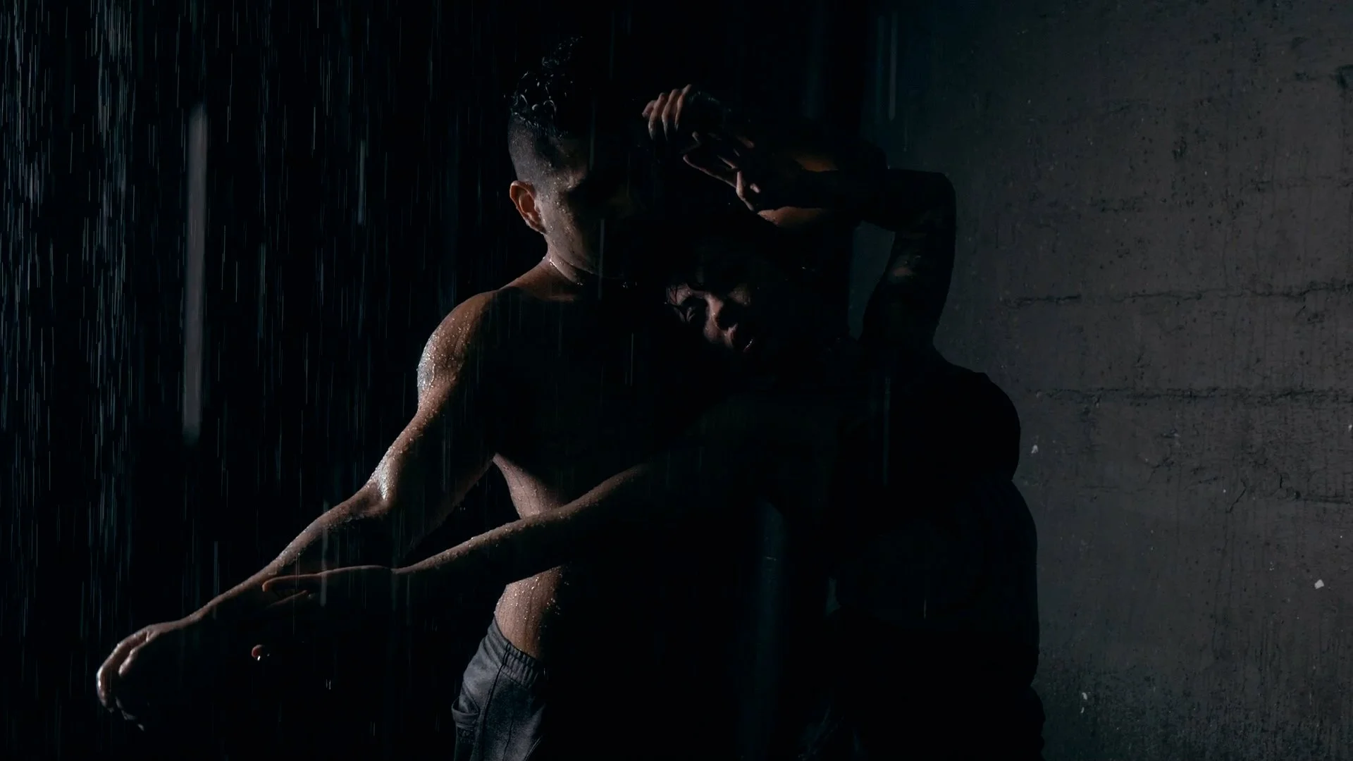 A person standing in the rain against a dark background, with water droplets visible on their skin and clothing.