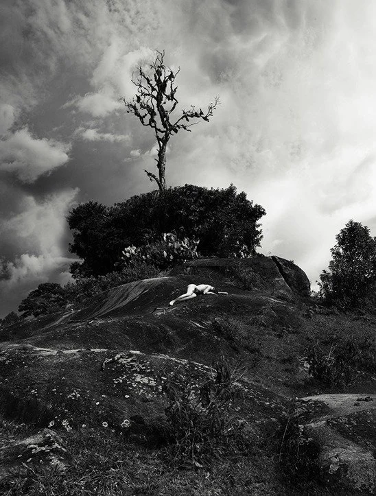 A person lying face down on a large rock formation outdoors, with a bare tree and other trees in the background under a cloudy sky, black and white photograph.