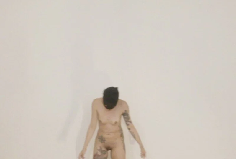Nude person with dark hair and tattoos standing against a plain white wall, head bowed and arms hanging down.