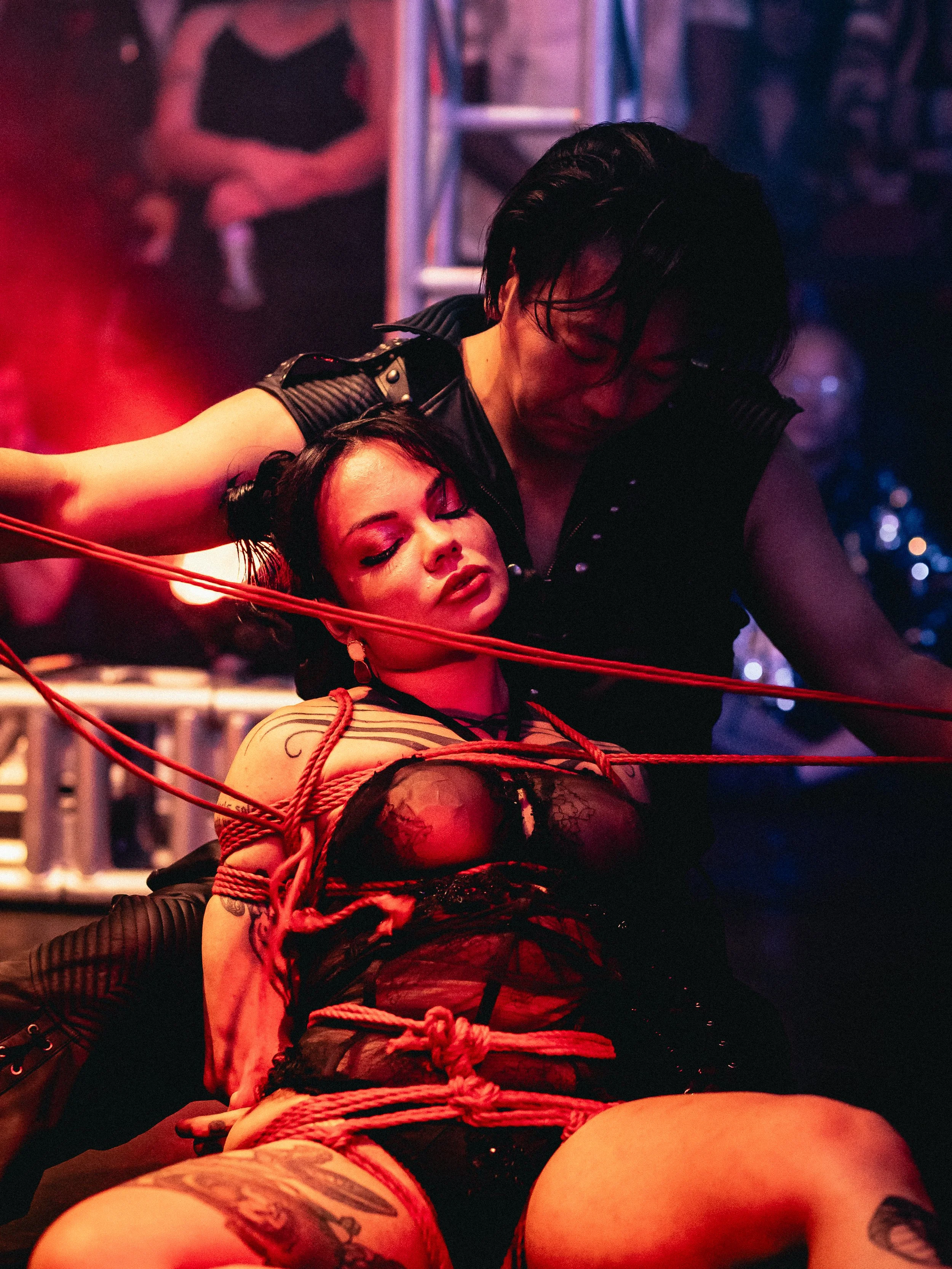 Two women in a dimly lit room with red lighting; one woman with short black hair, dressed in black, is leaning over another woman with long dark hair, who is tied with red ropes and dressed in black lace clothing. The woman with ties has her eyes clo