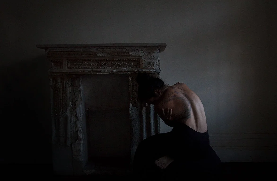 A person with tattoos sitting on the floor, hunched over with their head in their hands, next to an old, ornate fireplace in a dimly lit room.
