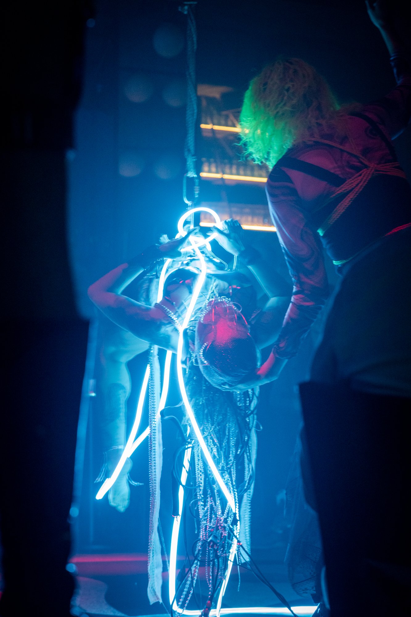 A person with neon green hair and dark clothing is interacting with a woman with neon blue lights or LED accessories in a dark, club-like environment with blue and purple lighting.