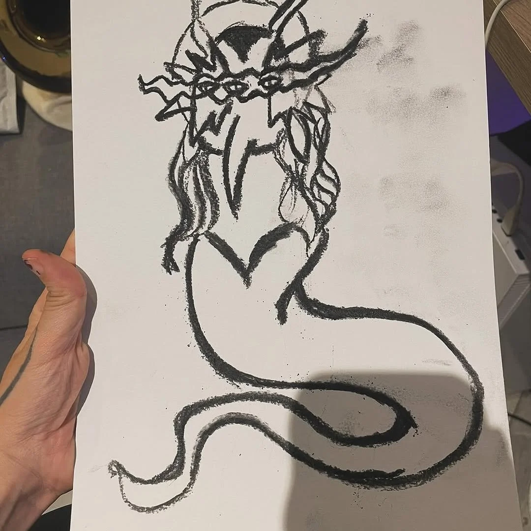 A black crayon drawing of a mythical creature with the head of a lion, multiple horns, and a long, curving serpent-like tail. The creature has a fierce expression and flowing mane.