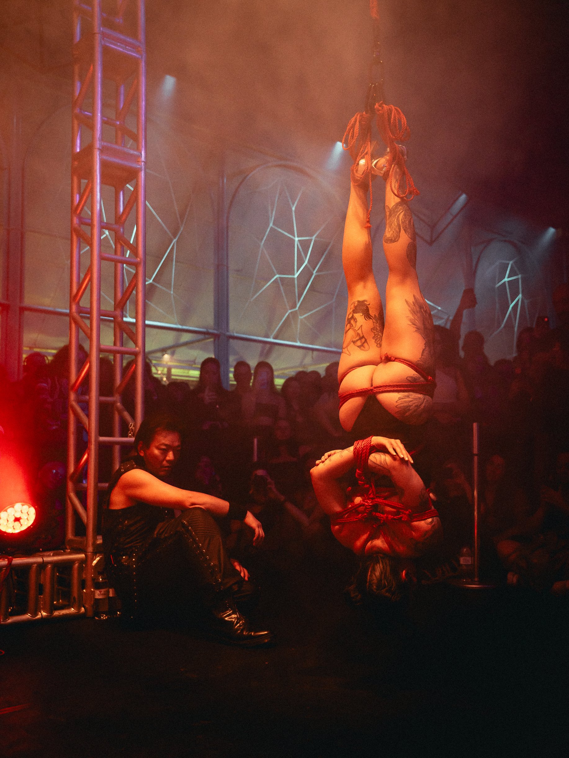 A tattooed woman performing an aerial silk routine on stage at a nightclub, with a seated person watching nearby and an audience in the background under colorful lights.