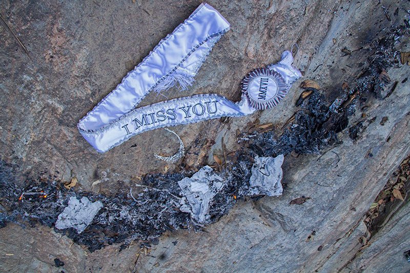 A memorial on a rock with charred wood and ashes, featuring a white ribbon and a rosette with a message that says 'I miss you'.