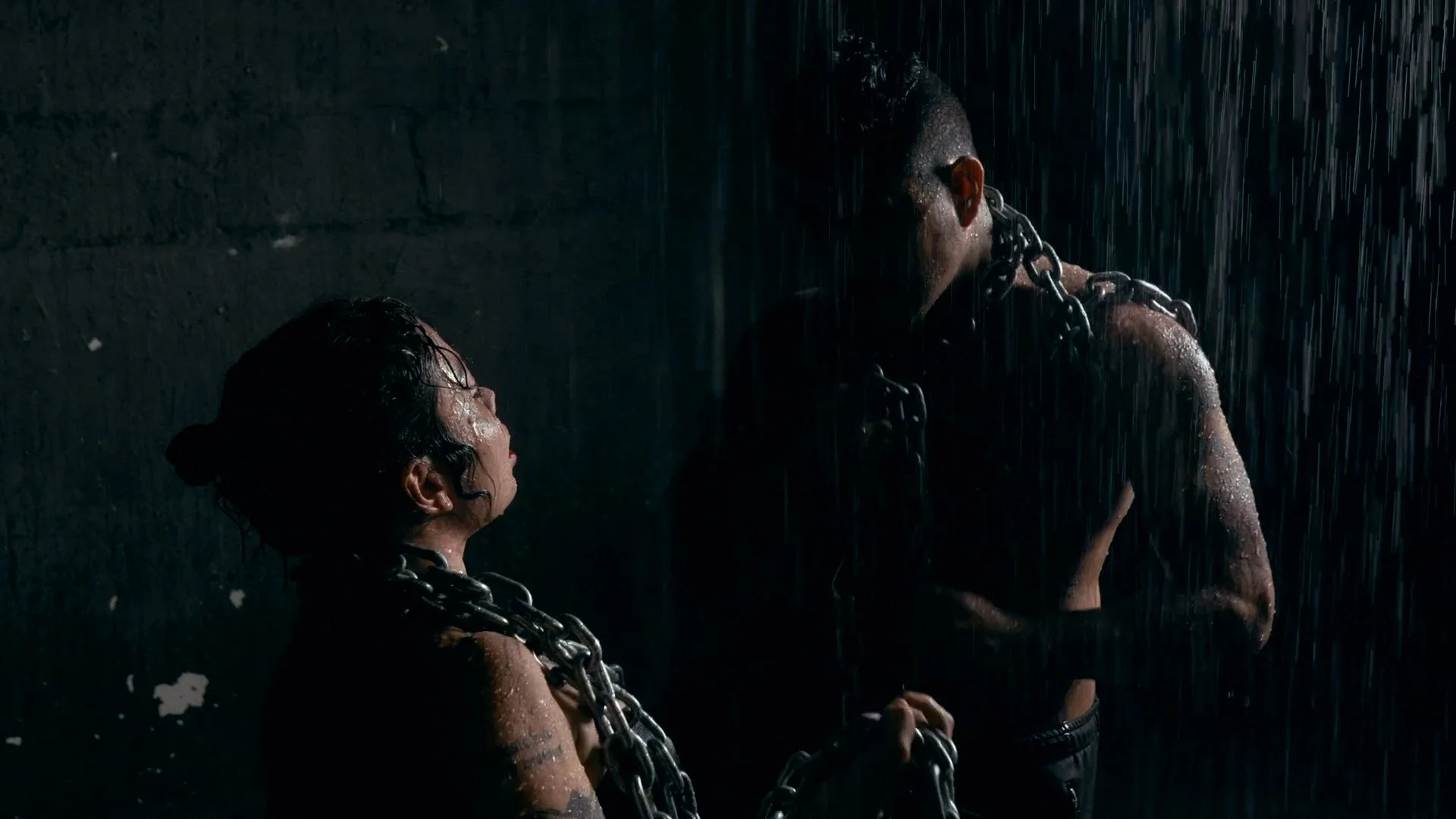 Two people wrapped in metal chains standing in the rain under a dark wall.