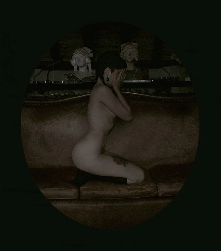 A woman sitting nude on a vintage sofa with her hands covering her face, in a dimly lit room with sculptures in the background.
