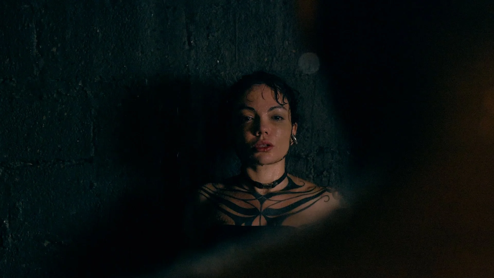 A woman with short wet hair and tattoos on her chest, lying against a dark brick wall at night, illuminated by a dim light.