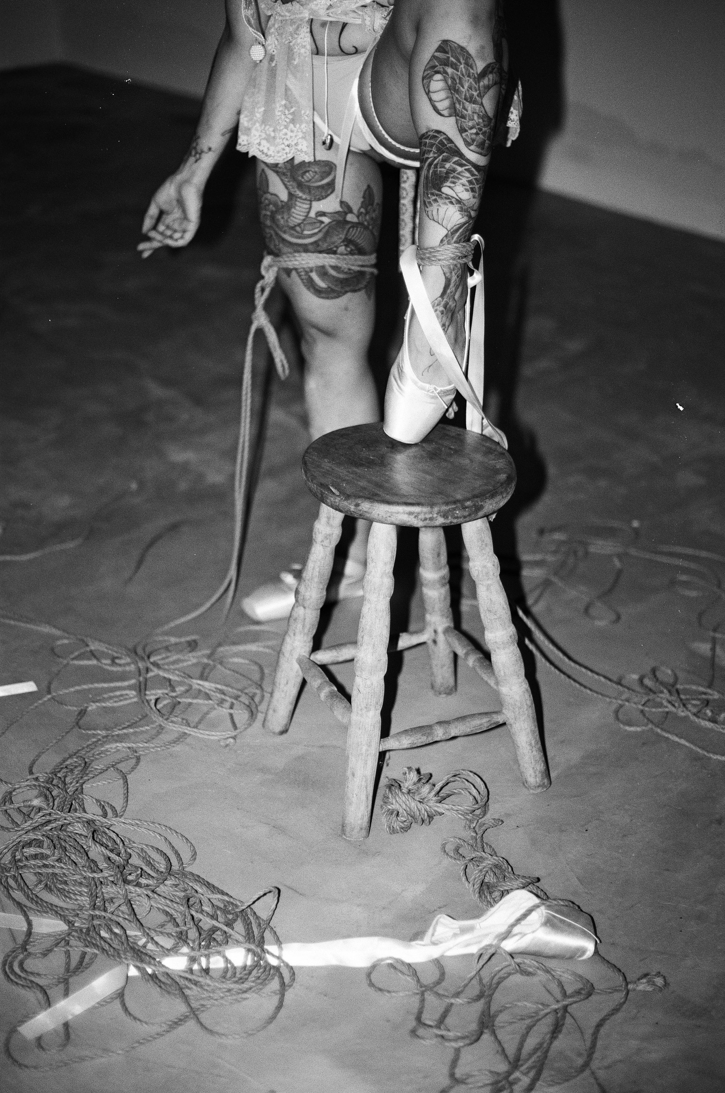 Tattooed woman in lingerie stands behind a wooden stool, with ropes and a ballet pointe shoe on the floor and on the stool.