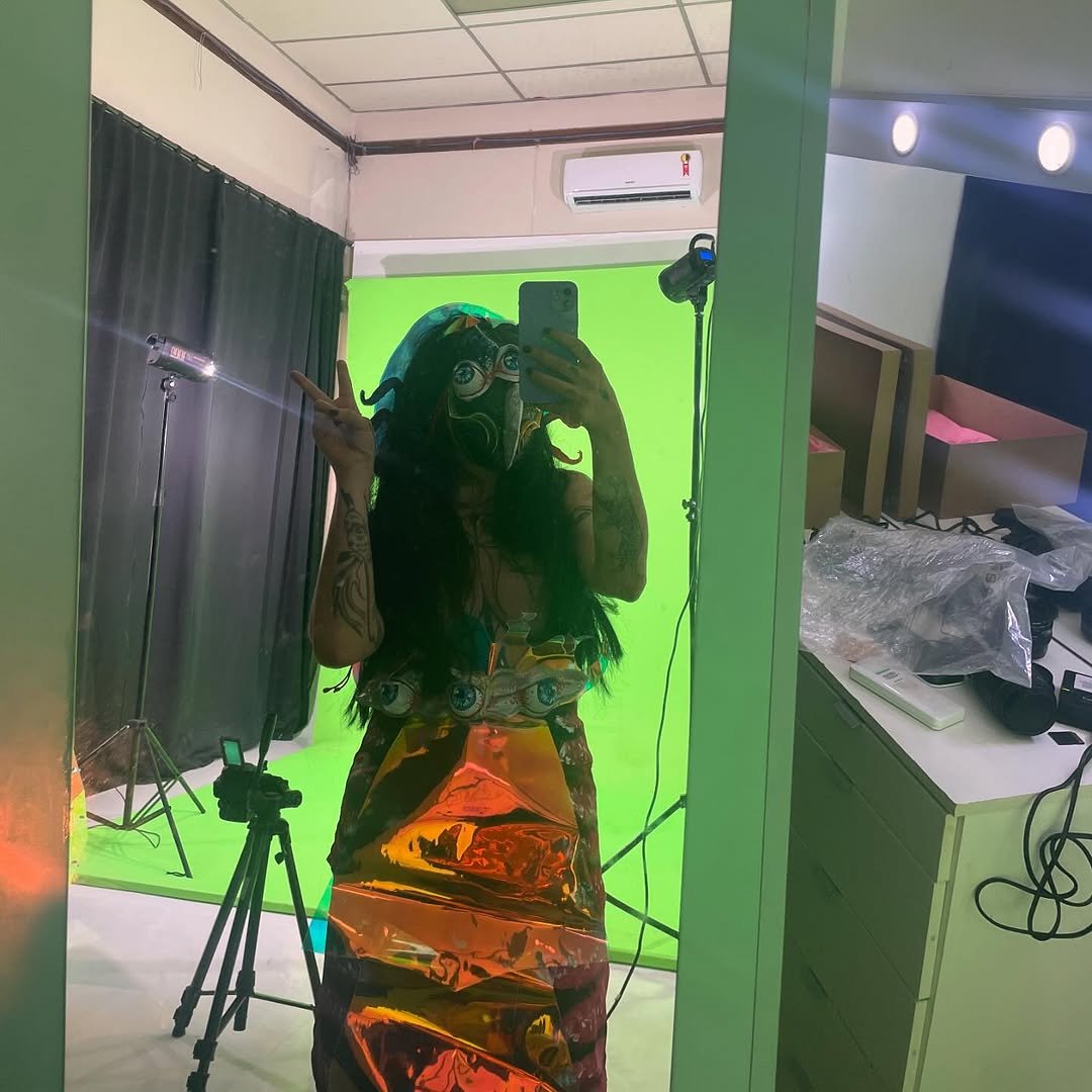 Person taking a mirror selfie in a studio with a green screen background, wearing a colorful mask and a dress with eye patterns, making a peace sign with their hand.