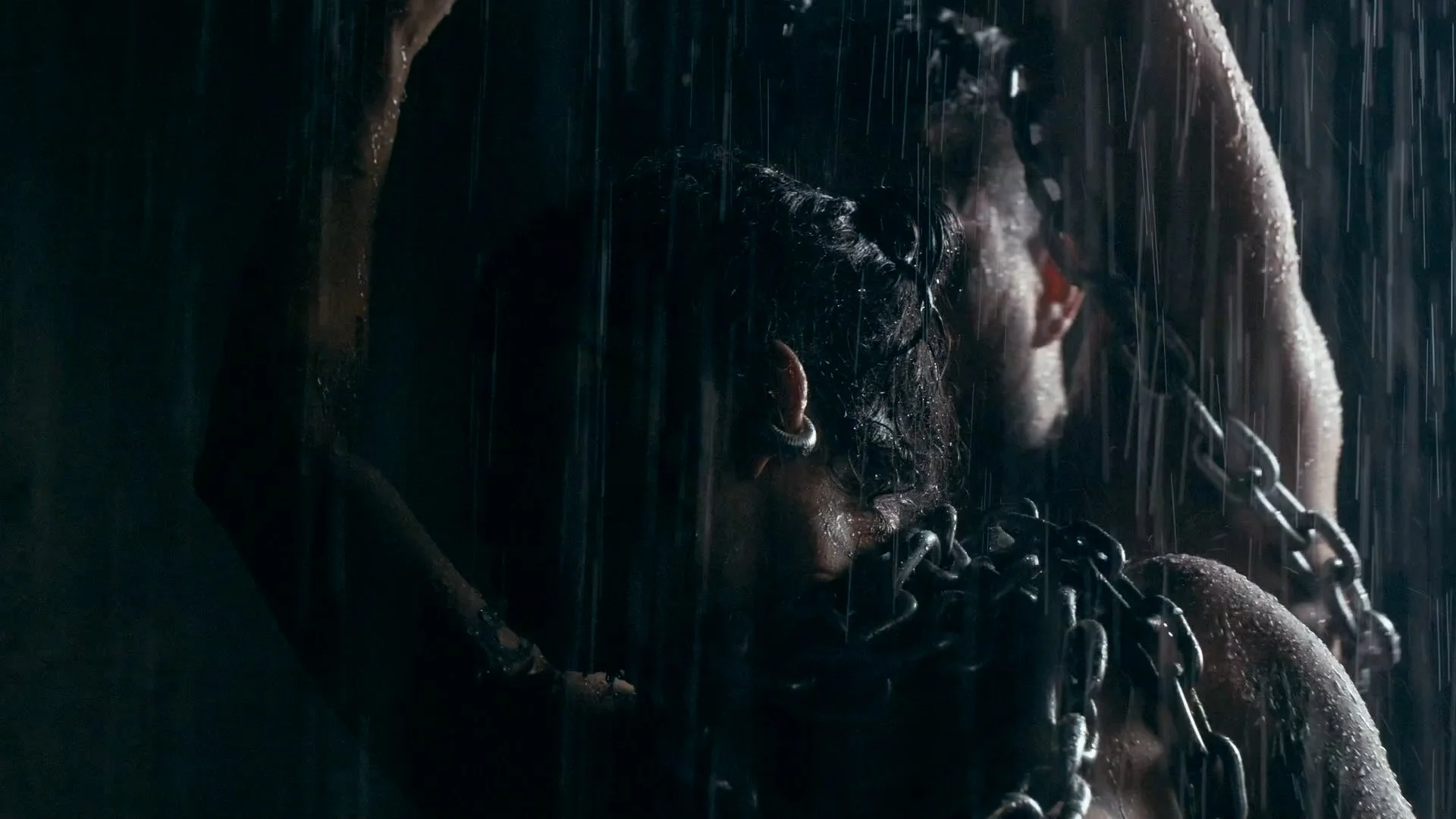 Close-up scene of a person in the rain with chains around their neck and shoulders, head down, dark and moody lighting.