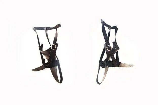 Two black harnesses with adjustable straps and metal buckles hanging against a white background.