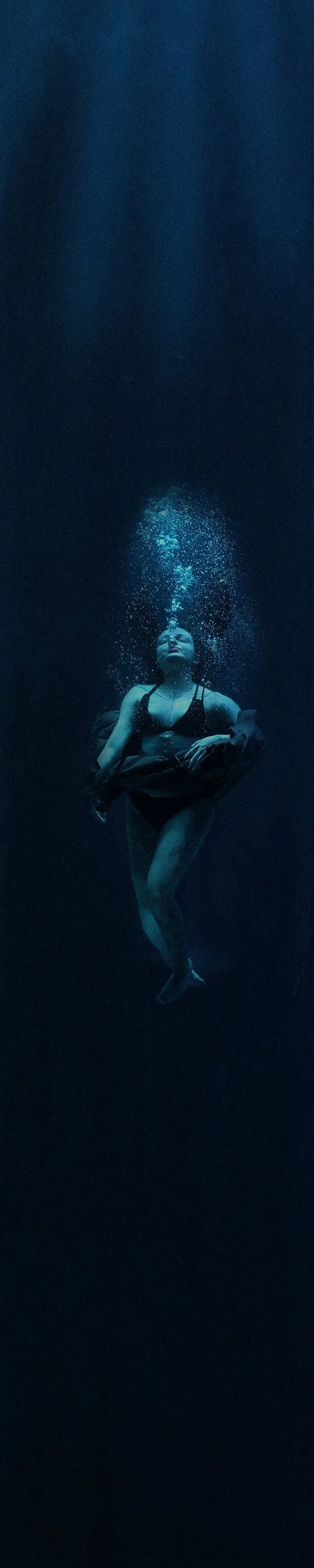 A woman in a black top is underwater, floating with her eyes closed and bubbles rising above her head.