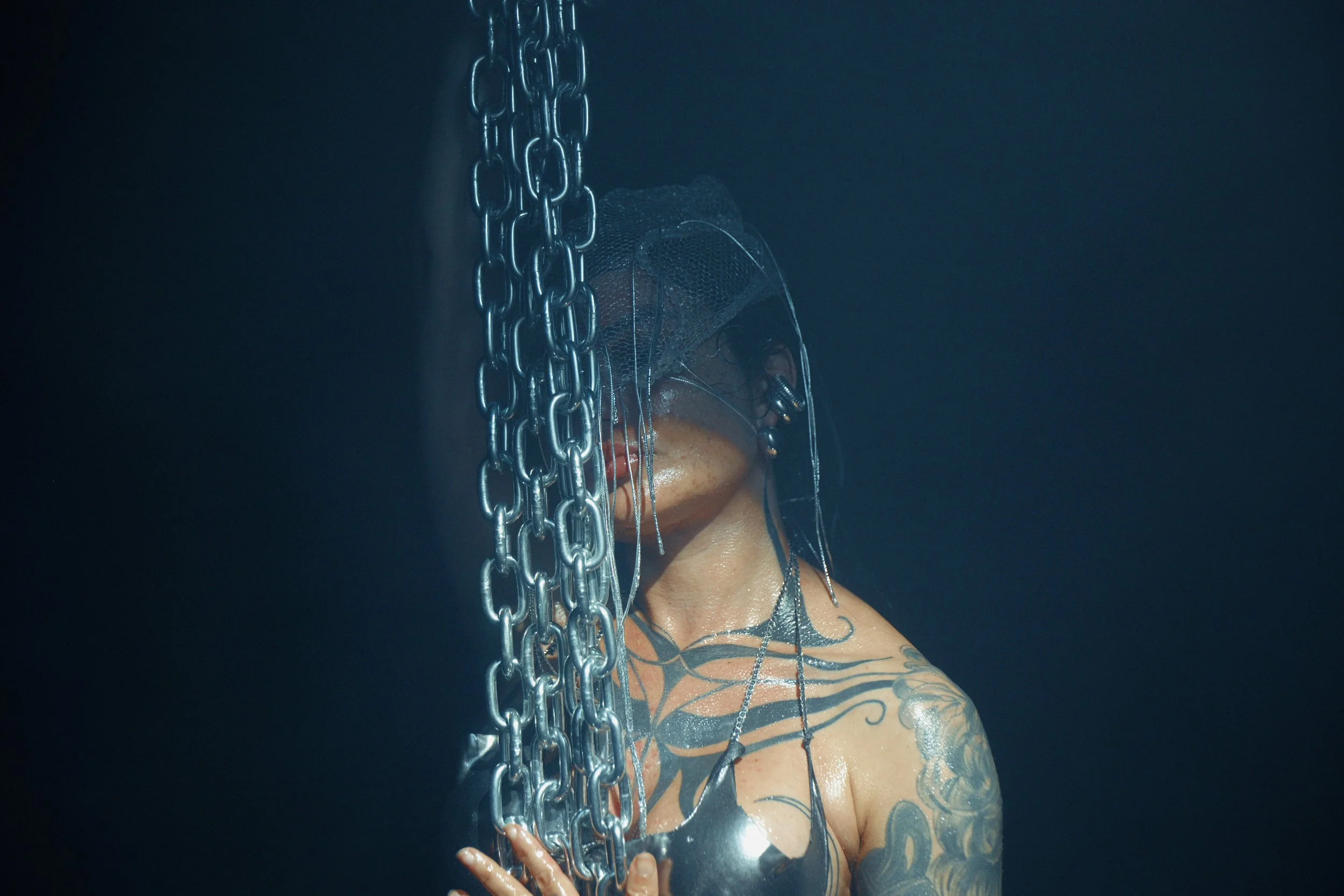 A woman with tattoos on her shoulder and arm holding onto a large chain, her face partially covered with reflective material and hair partially visible, against a dark background.
