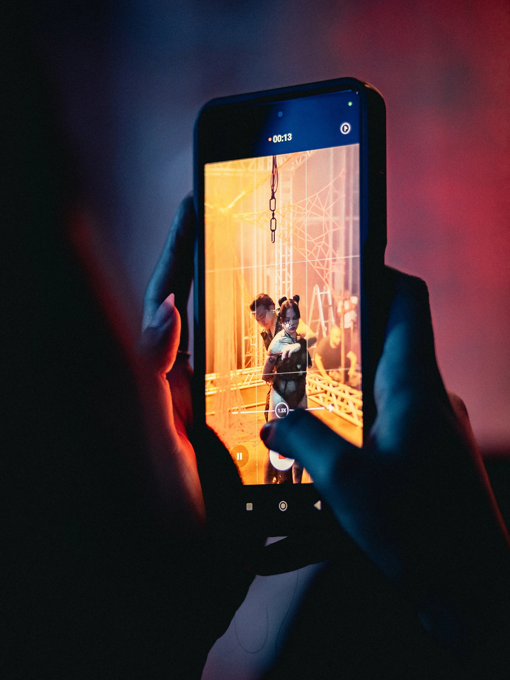 Person recording a video of two others taking a selfie in a dimly lit, Halloween-themed setting with orange lighting and cobweb decorations.