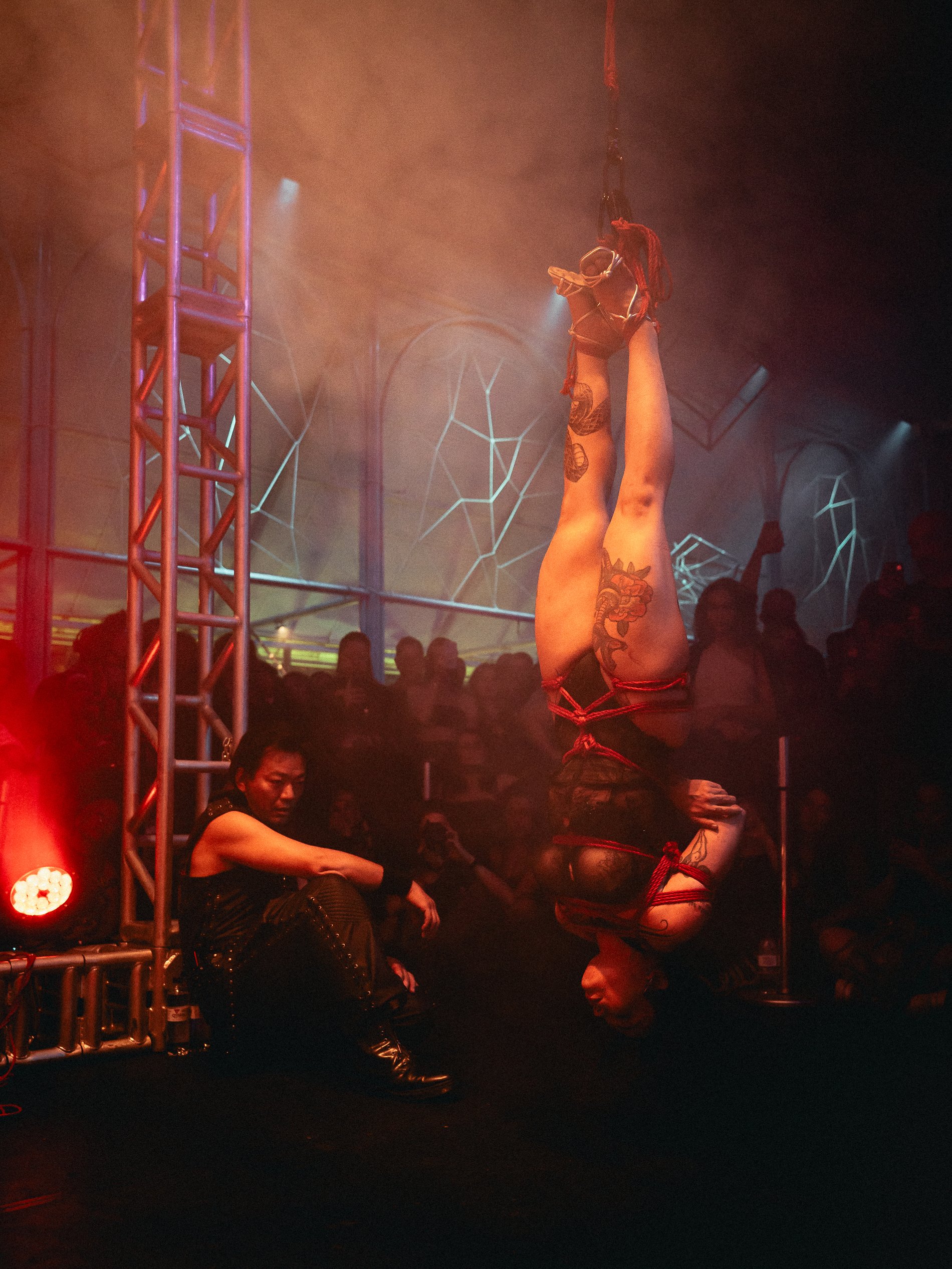 A woman doing an aerial acrobatic performance upside down, attached to a swing in a dimly lit, vibrant nightclub or performance venue filled with spectators.