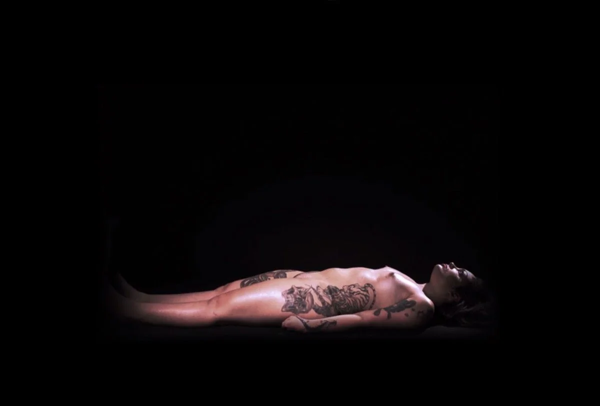 A nude woman with tattoos lying on her back against a black background.