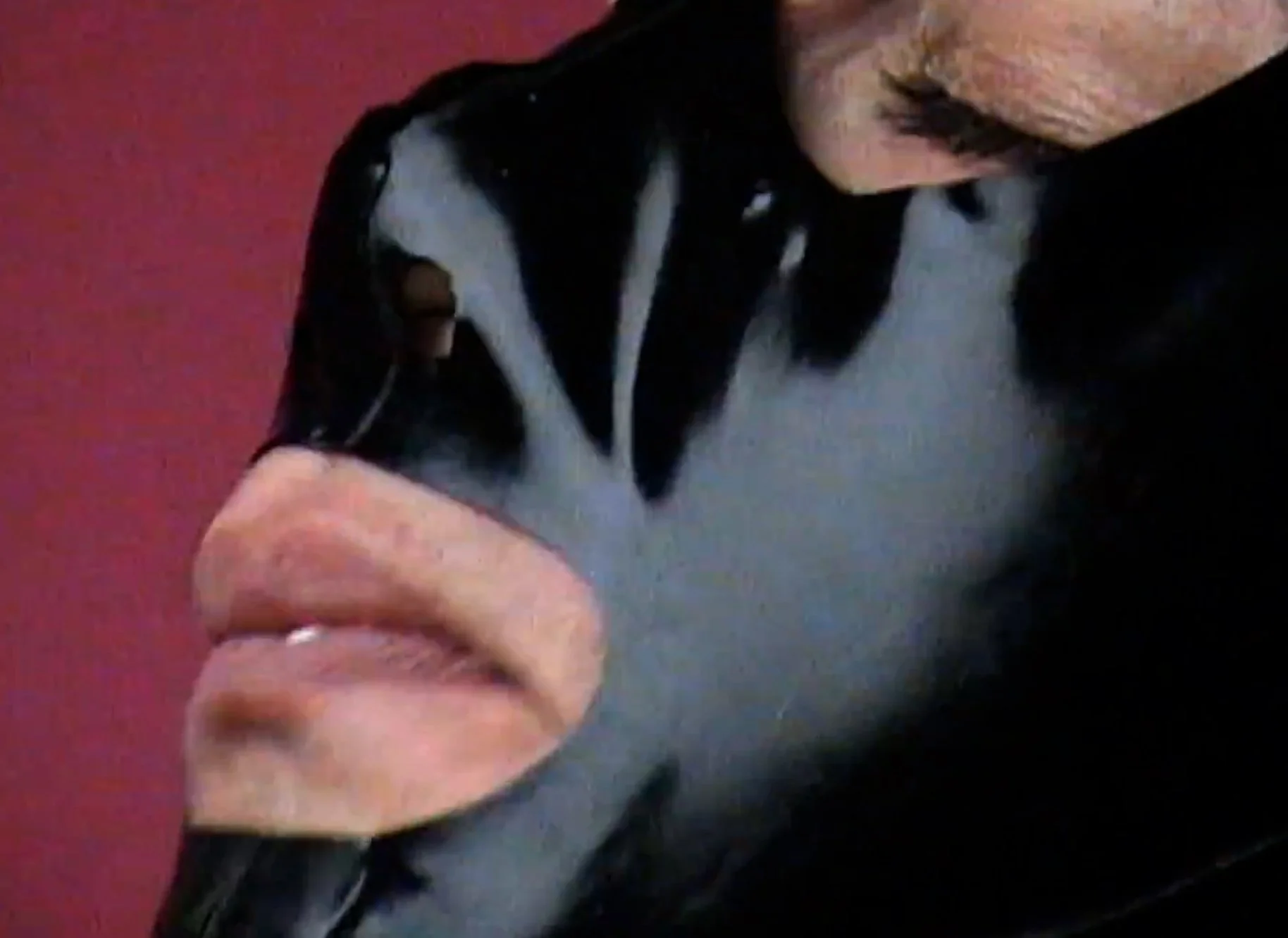 Close-up of a person with black and gray shirt, showing their chest, chin, lips, and part of their face with a reddish background.