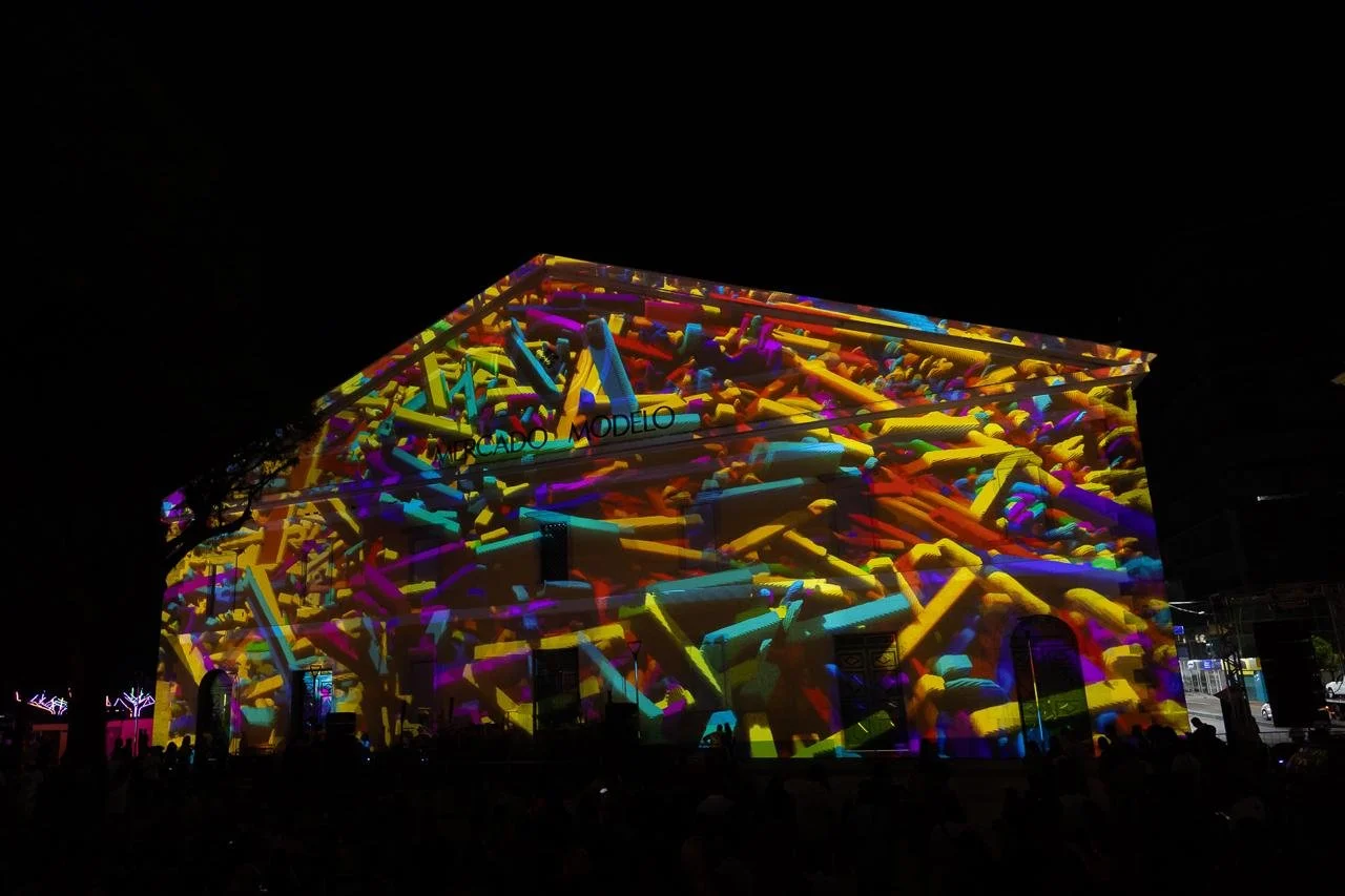 Nighttime projection mapping on the side of a large building, displaying colorful, abstract shapes in vibrant yellow, pink, purple, and blue.