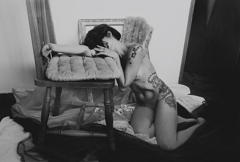A person with tattoos kneeling on the floor, resting their head and arms on a stool, with a disheveled bed and blanket in the background.