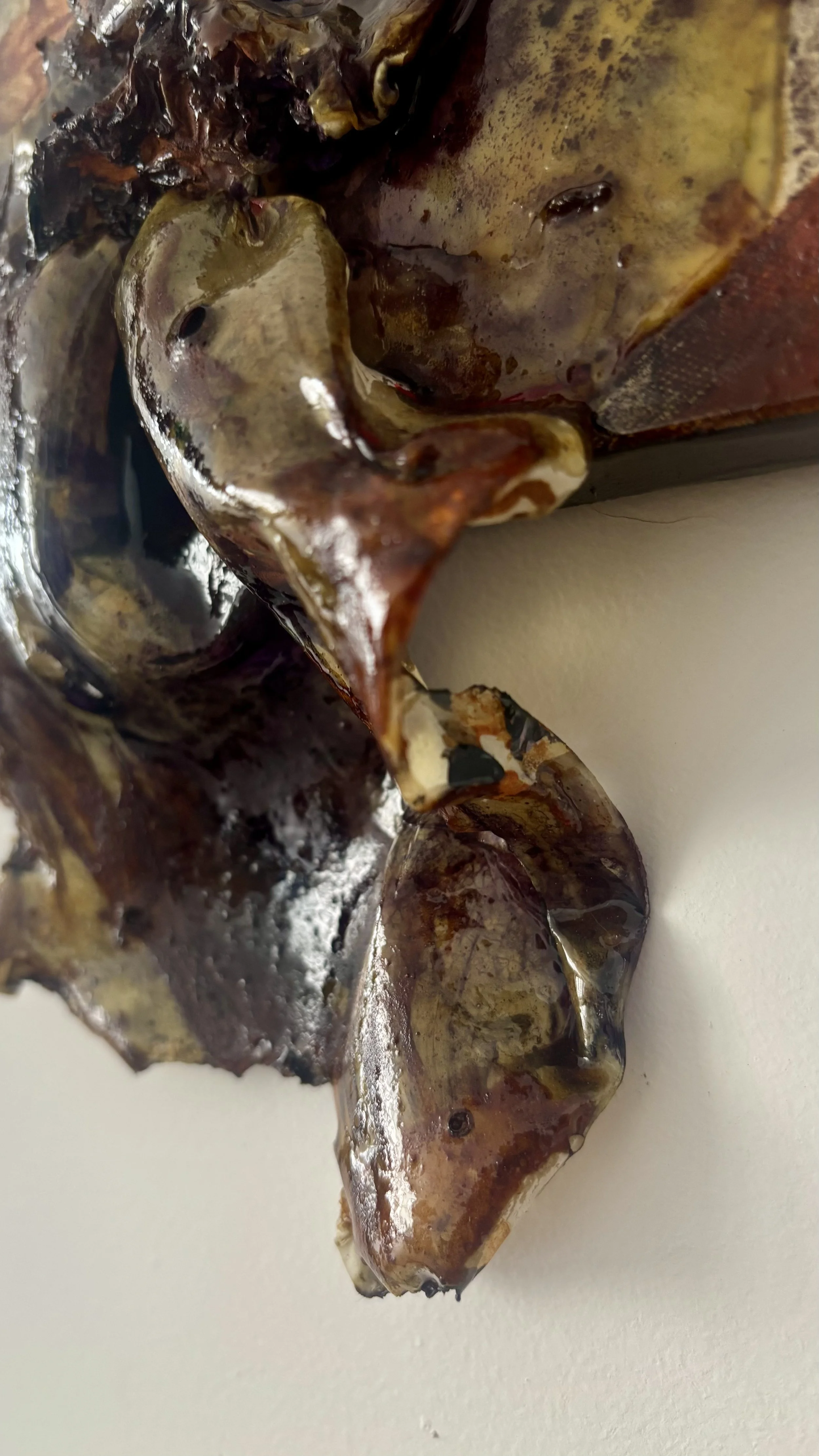 Close-up of burnished, melted, and partially charred plastic or metal object with irregular shapes and textures, predominantly brown and black with some yellowish areas, placed against a white surface.