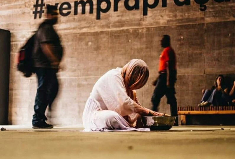 A woman wearing a headscarf kneeling on the floor, feeding or giving something to a dog in a bowl. In the background, there are blurred figures of people walking and sitting against a brick wall with the hashtag '#remkraps' partially visible.