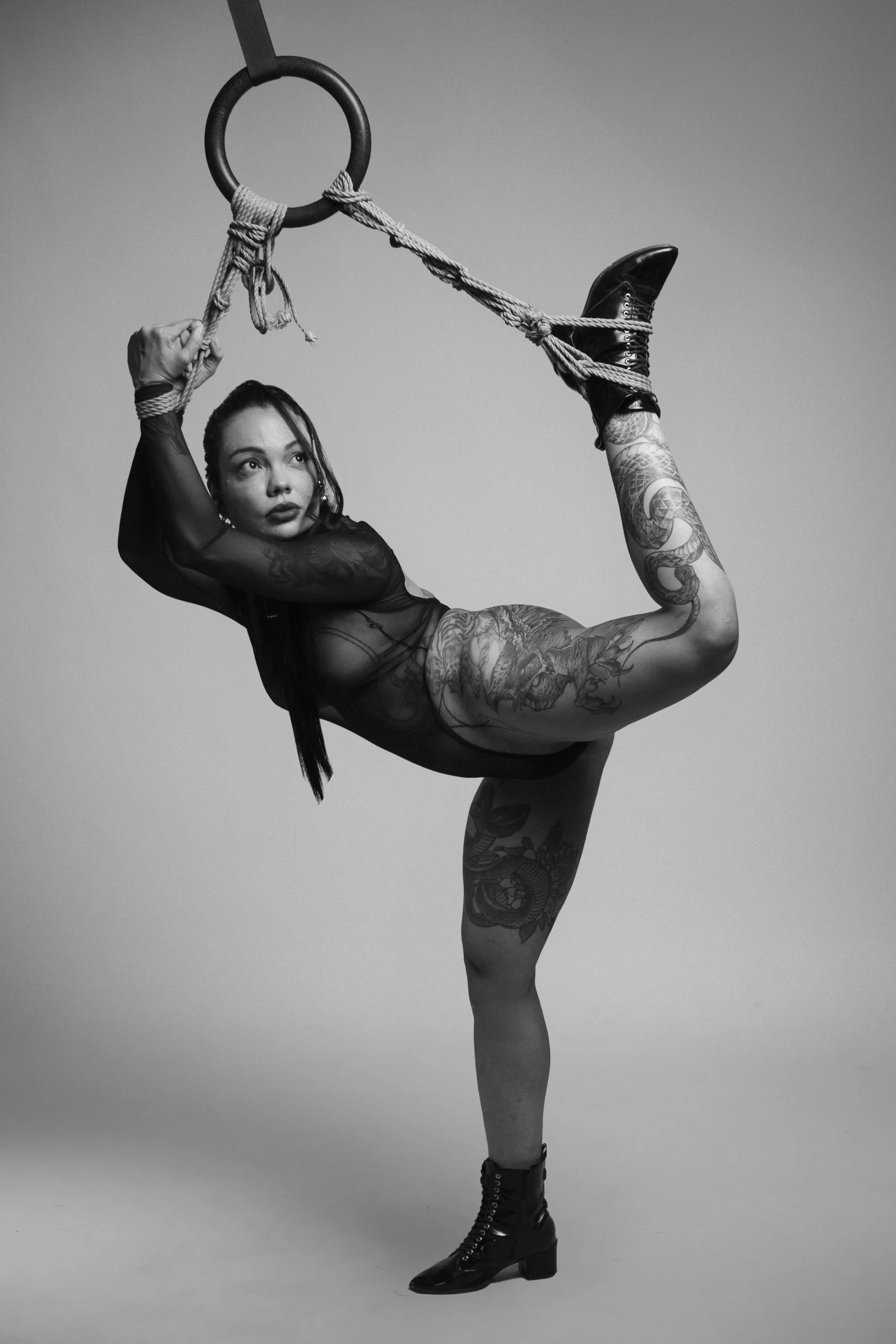 A woman with tattoos in a black mesh outfit and black ankle boots performs a contortion pose on an aerial ring with a rope, against a plain background.