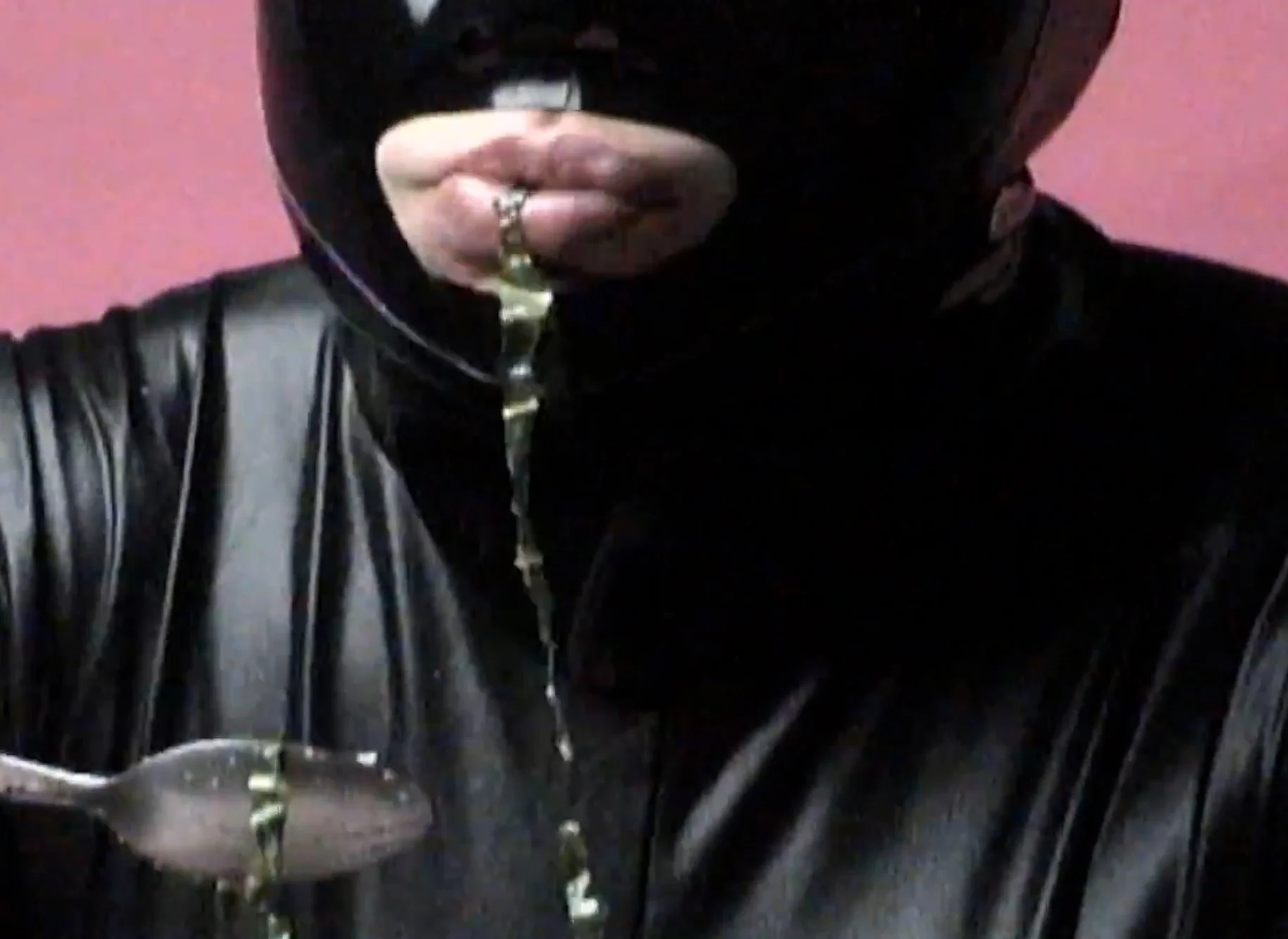 A person wearing a black ski mask, black leather jacket, and holding a metal fork with a gold handle, with pink background.