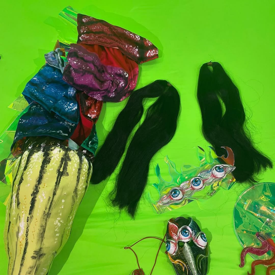 A collection of colorful, shiny packaging and accessories, including artificial hair, candy, and a toy with eyes, laid out on a bright green surface.